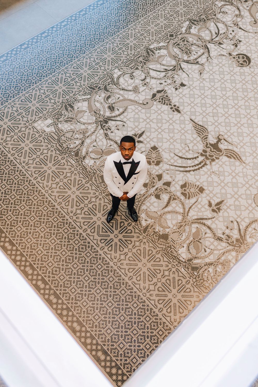 Aerial view of a groom in a white tuxedo standing on an ornate patterned tile floor.
