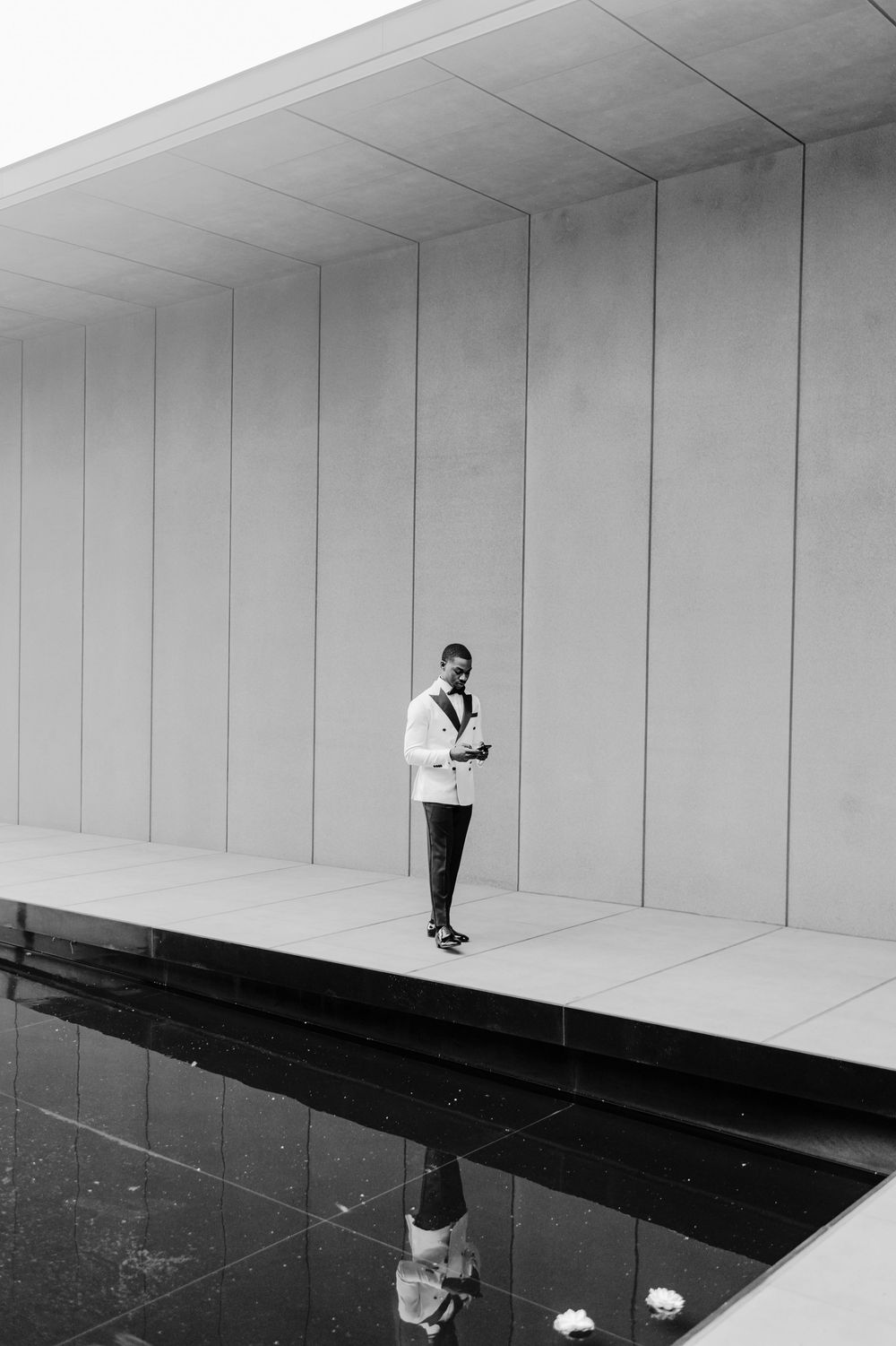 Black and white photo of a man in a white blazer standing by a reflective pool against a modern concrete wall.