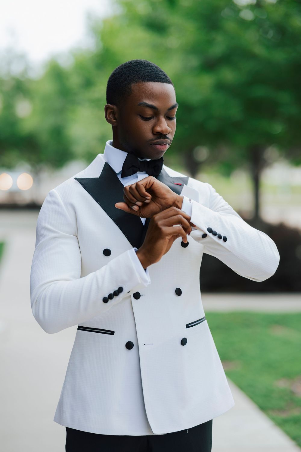 Young Black man in white tuxedo jacket with black lapels and bow tie adjusting cufflinks outdoors.