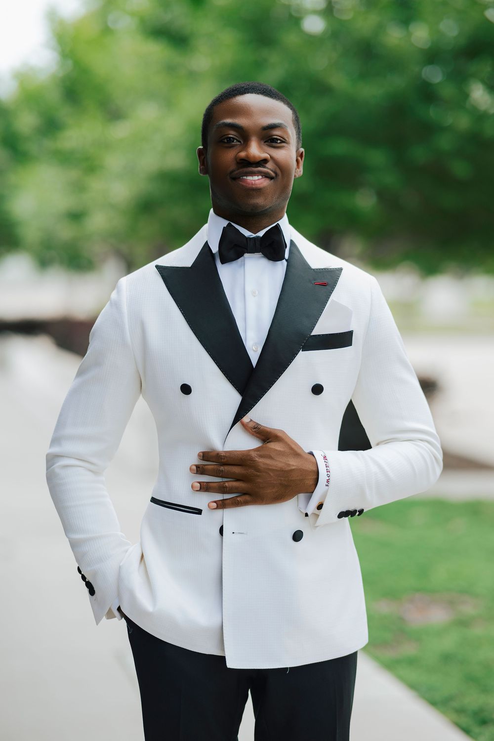 Smiling man in white double-breasted tuxedo jacket with black lapels and bow tie posing outdoors.