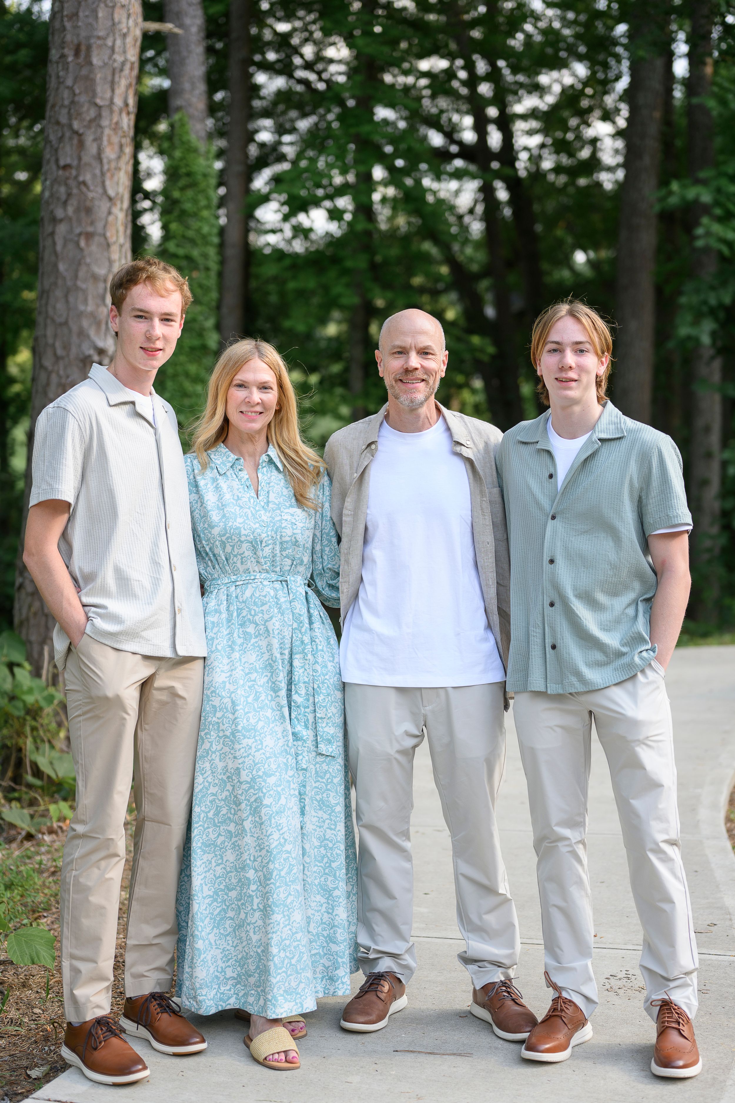 Family of four smiling outdoors among tall trees, wearing casual summer outfits in neutral and pastel tones.