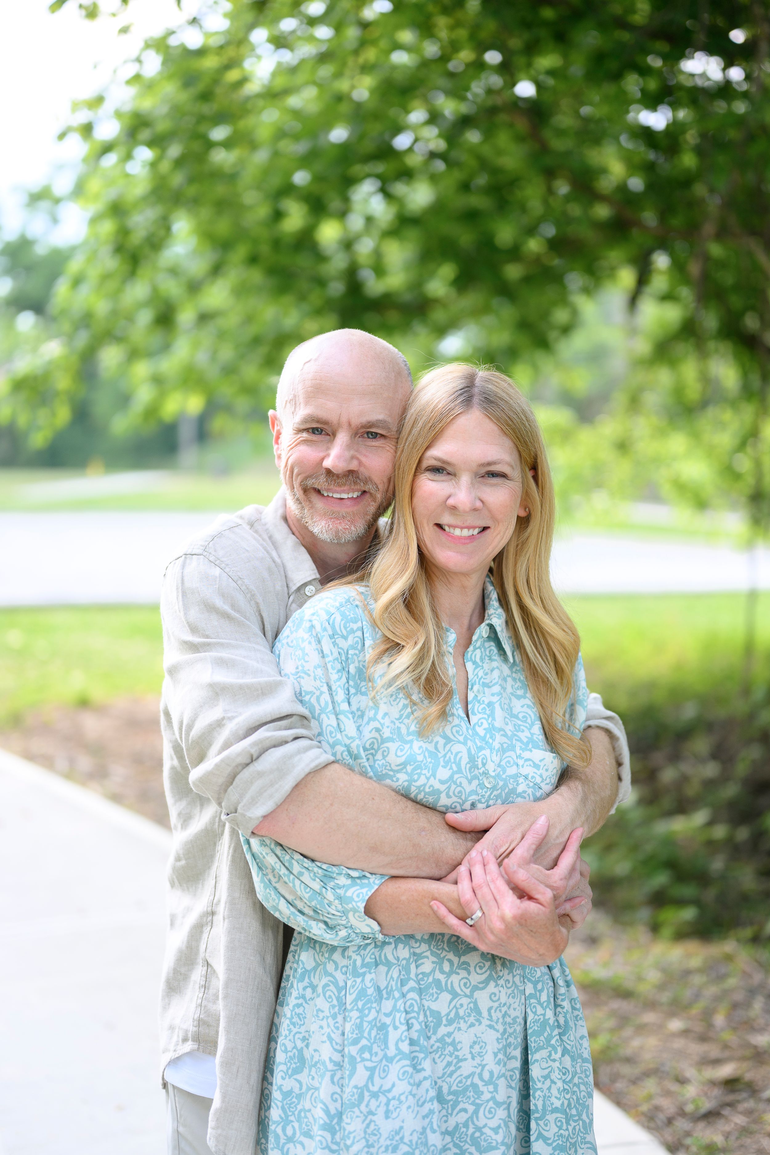 A smiling couple embraces outdoors on a sunny day, with lush green trees in the background.