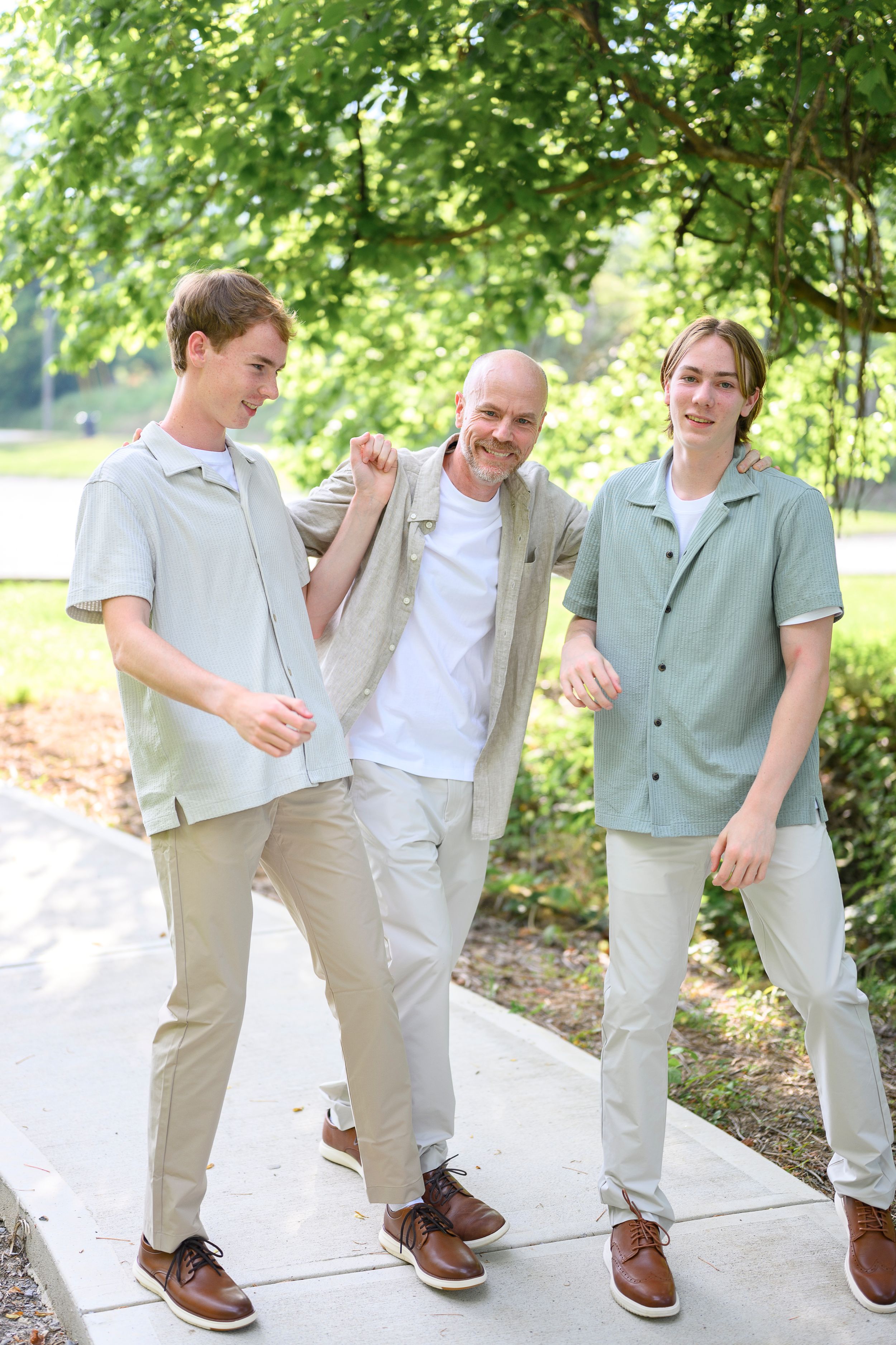 Three men in coordinating neutral outfits pose together on a sunny park path surrounded by green trees.