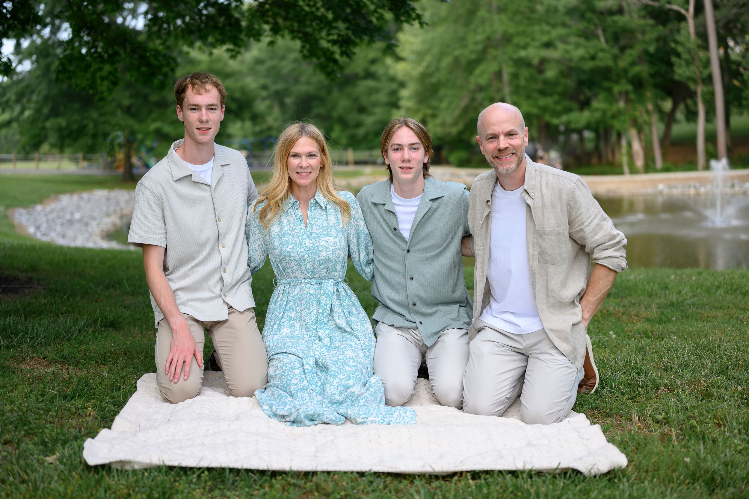 Family of four posing on a blanket in a lush green park near a pond on a sunny day.
