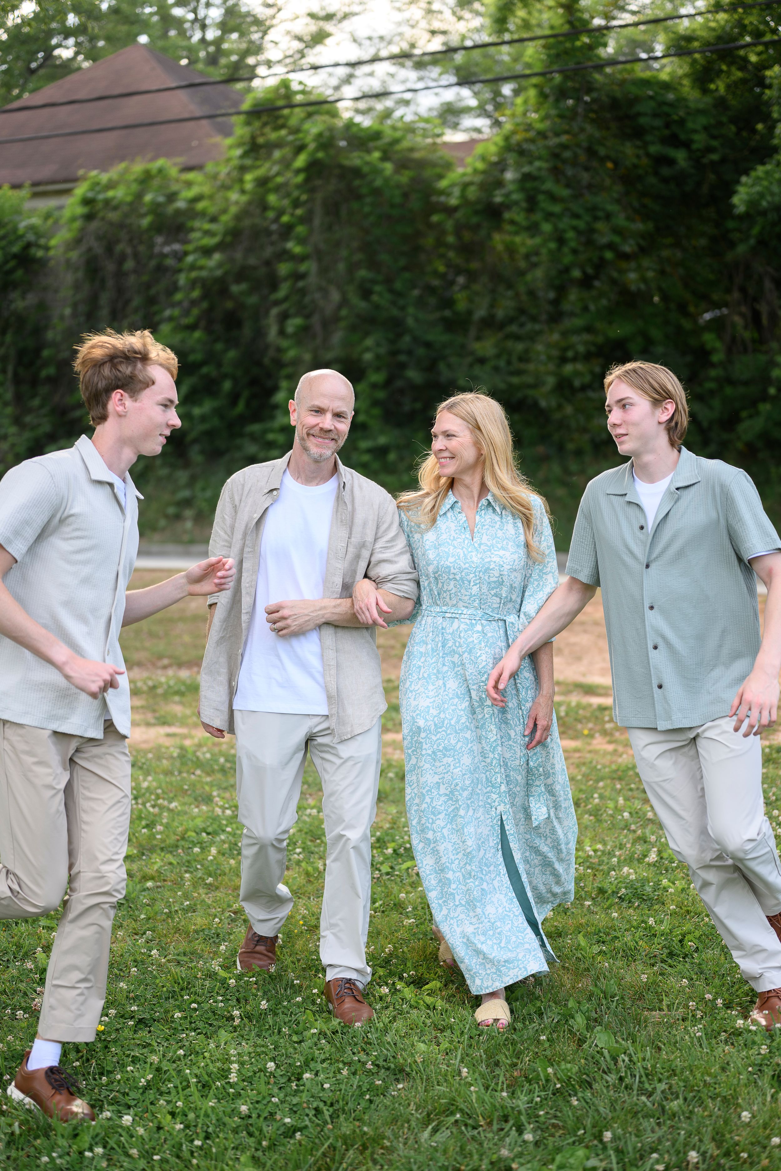 Family of four walks outdoors on green grass, smiling and conversing, dressed in casual summer attire near lush greenery.