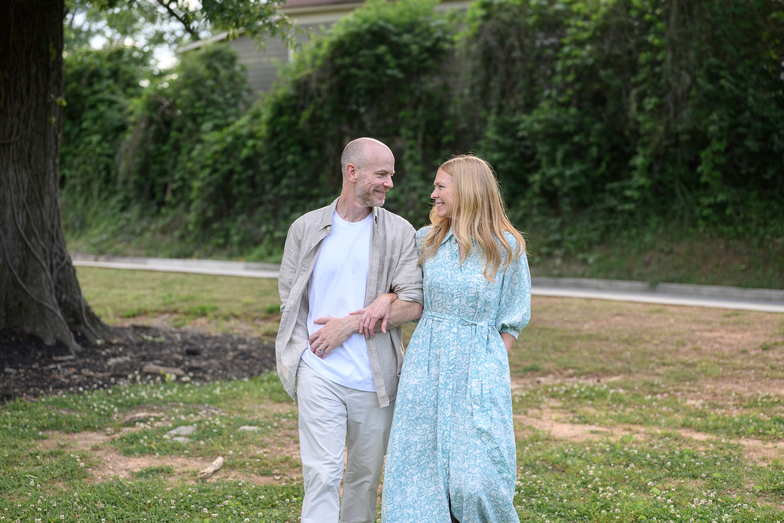 A smiling couple walks arm in arm through a lush green park, the woman in a floral blue dress and the man in casual beige attire.