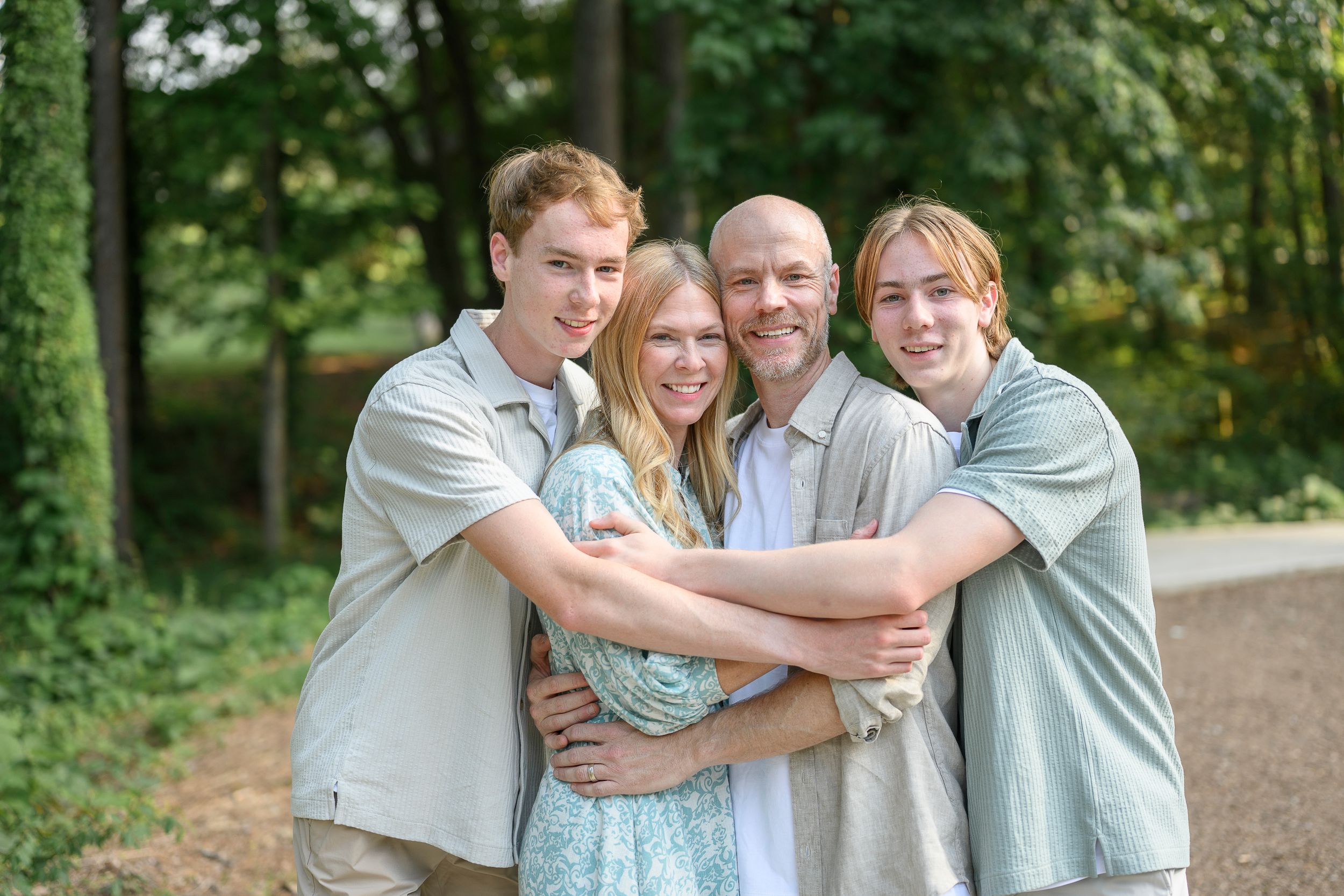 Family of four smiling and hugging outdoors on a path surrounded by lush green trees.
