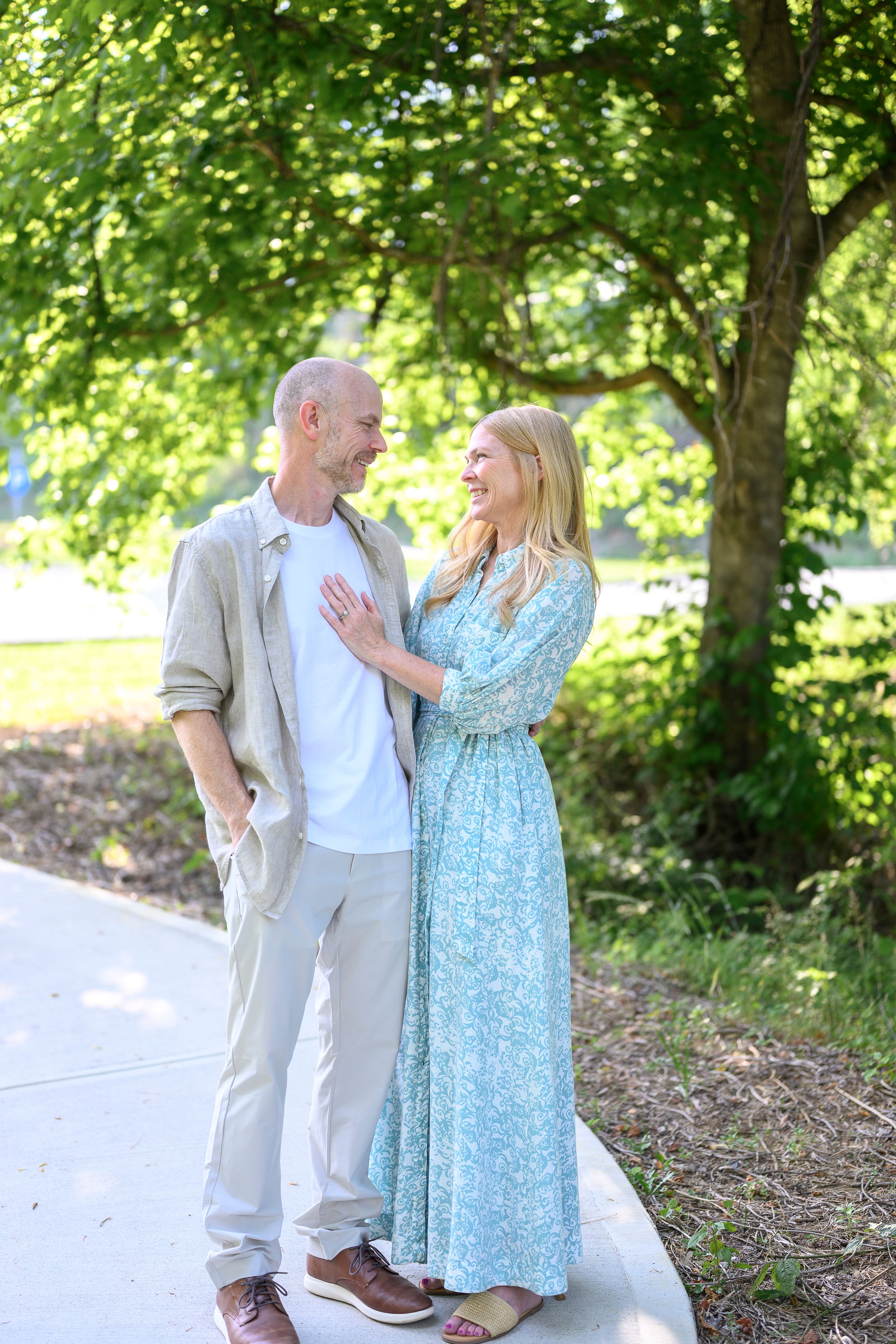 A smiling couple shares a tender moment on a sunny path surrounded by lush green trees in a park.