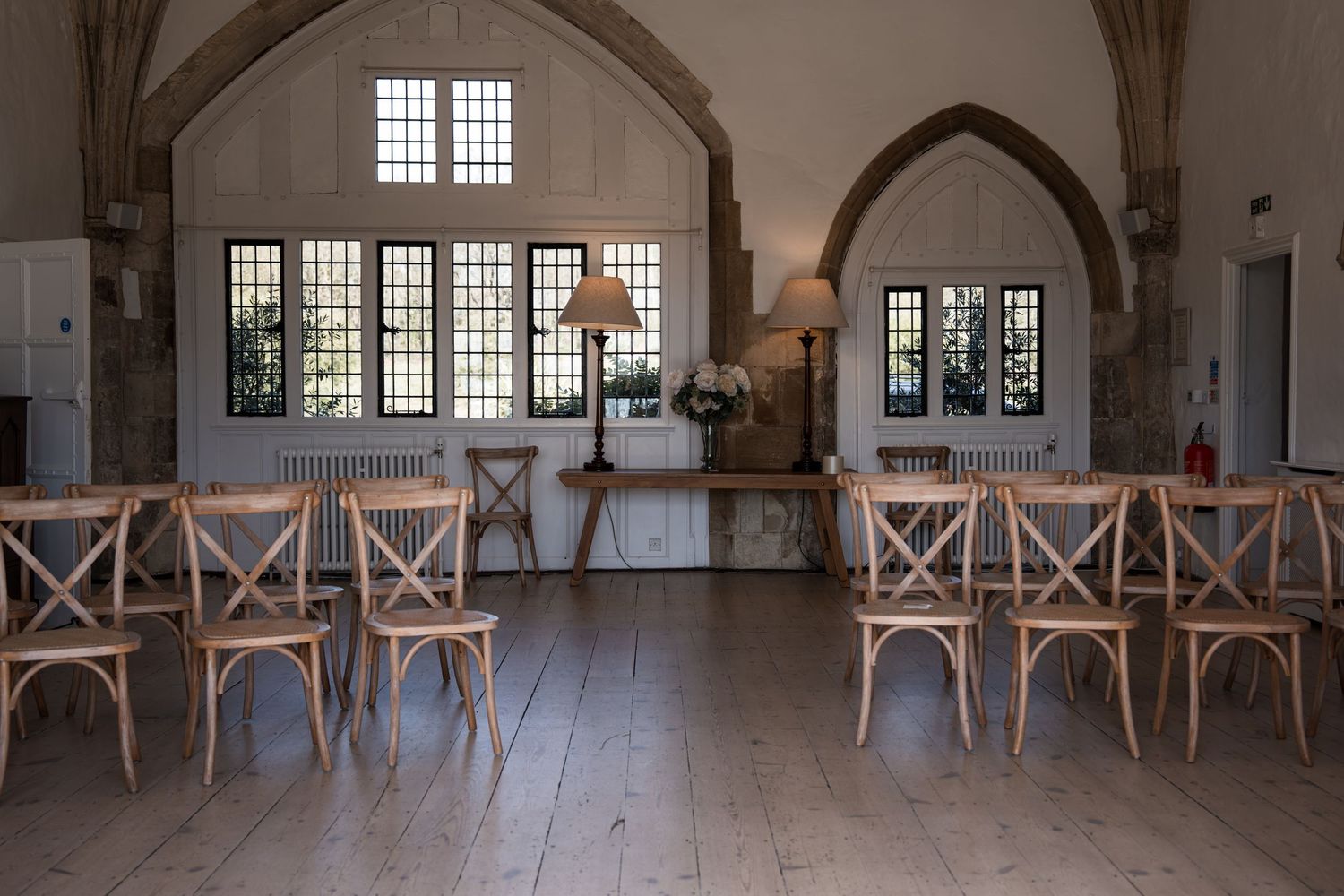 Rustic wedding ceremony room with wooden cross-back chairs and arched windows in historic venue.