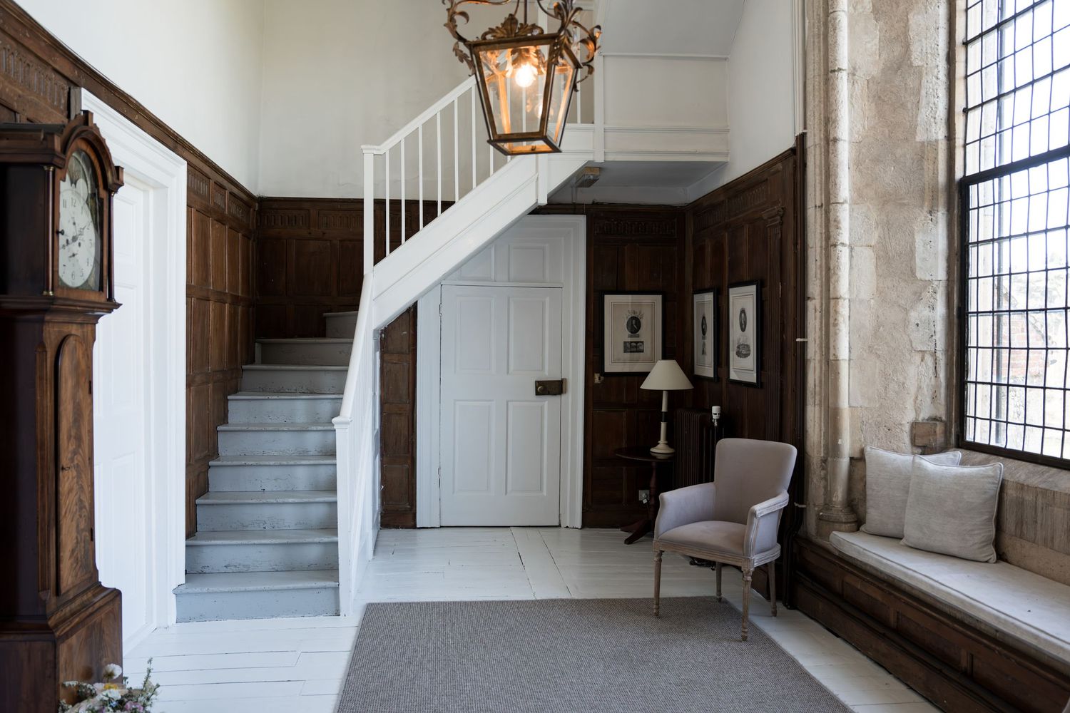Grand historic manor hallway with dark wood paneling, white staircase, lantern chandelier and window seat.