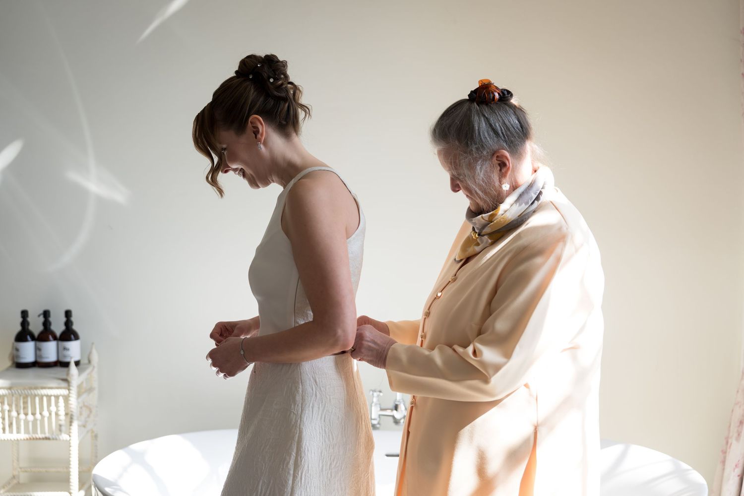 Older woman helps bride fasten white wedding dress in bright preparation room before ceremony.