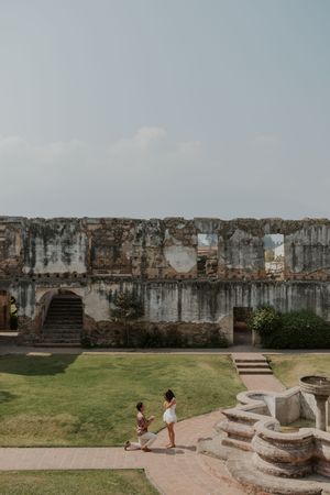 Couple sharing a romantic proposal moment at ancient stone ruins with lush green lawn under a hazy sky.