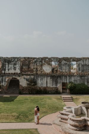 Woman exploring ancient stone ruins with lush green lawn, arched doorways, and historic weathered walls under a hazy sky.