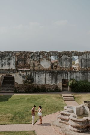 Couple holding hands at ancient stone ruins with lush green lawn, romantic engagement photo at historic landmark.