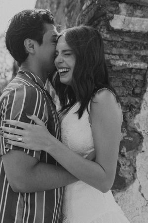 Black and white photo of a laughing couple embracing near a rustic stone wall, sharing a joyful romantic moment.