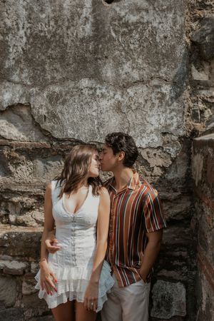 A young couple shares a romantic kiss against a rustic stone wall, wearing summer outfits.
