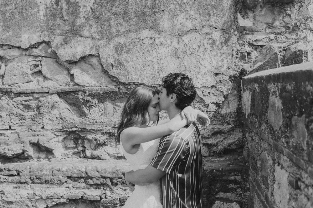Black and white photo of a couple sharing a romantic moment against a rustic stone wall.