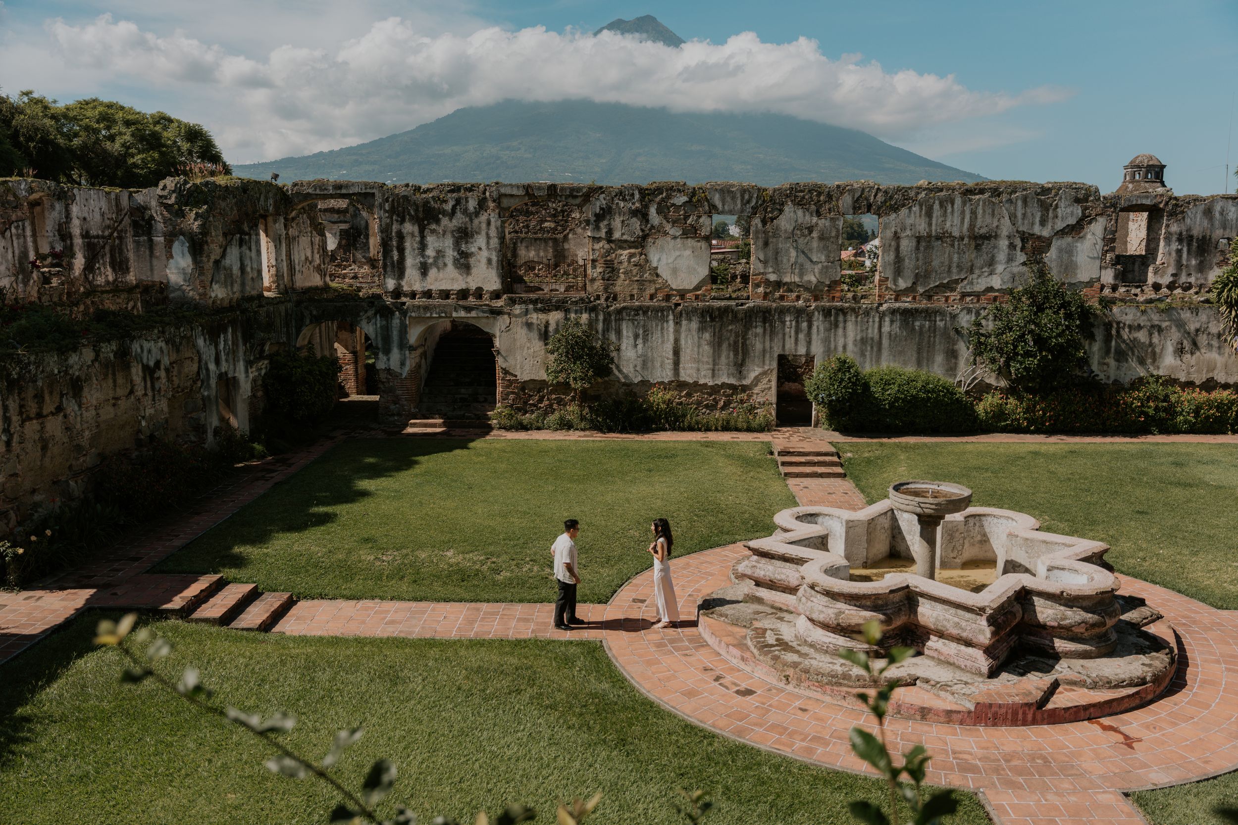 Couple standing in lush courtyard of ancient stone ruins with volcanic mountain backdrop.