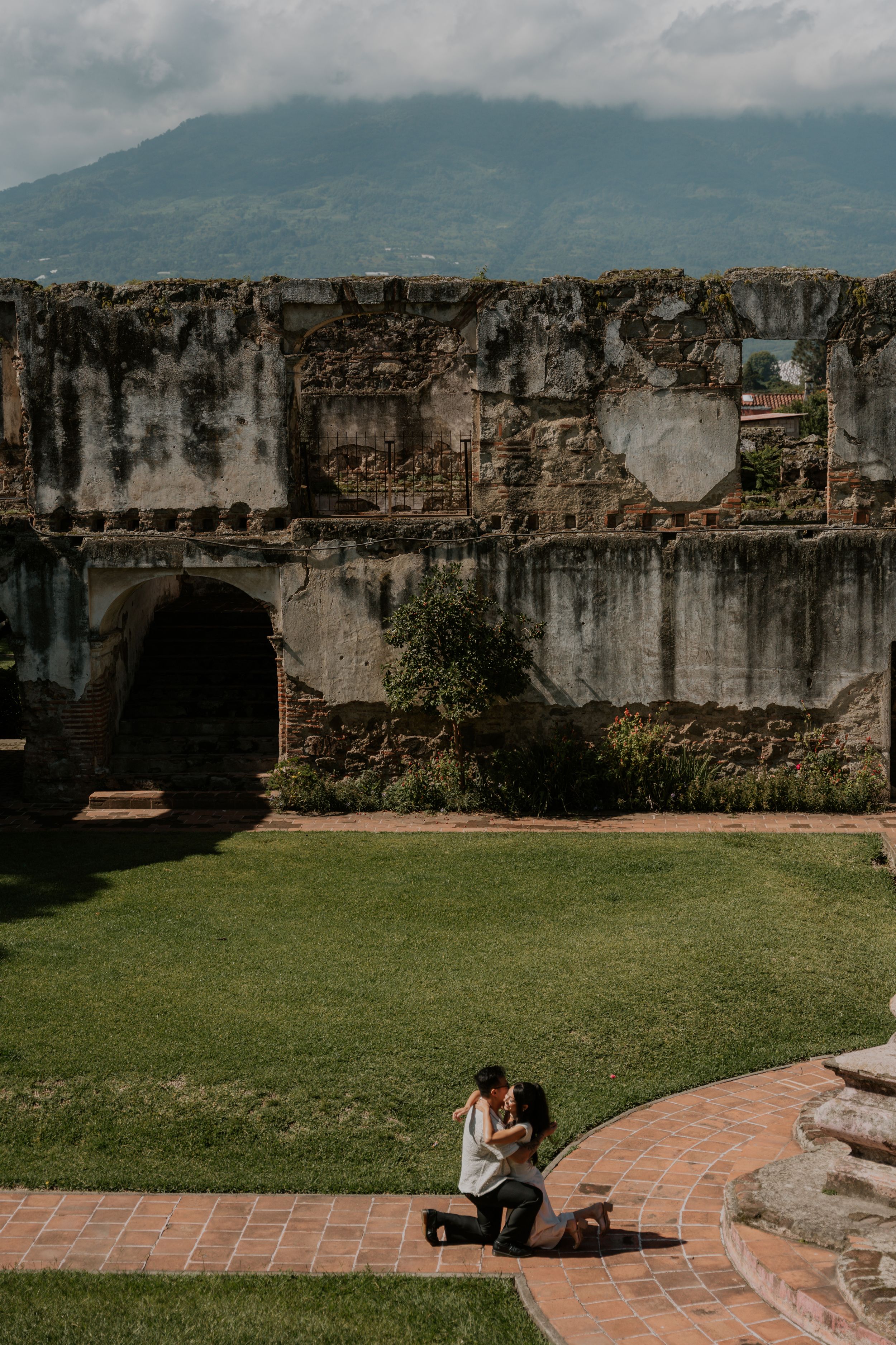 Couple sharing a romantic moment in front of ancient stone ruins with lush green lawn and mountain backdrop.