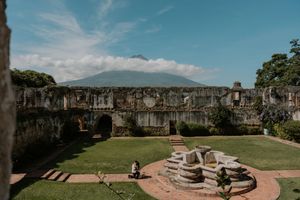 Ancient stone ruins with lush green courtyard and volcanic mountain backdrop under blue sky in Antigua, Guatemala.