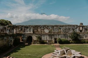 Ancient stone ruins with arched doorways and lush green lawn, with a volcano rising dramatically behind under a cloudy sky.