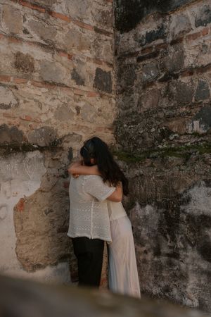 Couple embracing intimately in a rustic stone corner, woman in white lace top and flowing skirt.