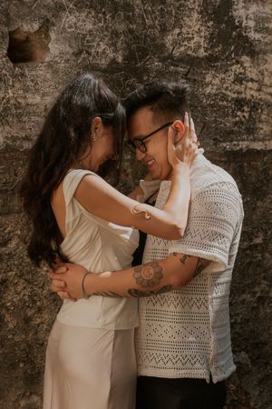 A couple shares a tender embrace against a rustic wall, the man wearing glasses and a crochet shirt, the woman in a white outfit.