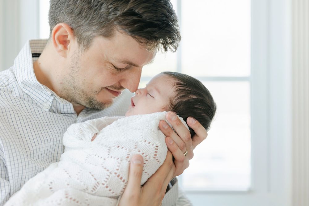 Father tenderly holds sleeping newborn baby wrapped in white knit blanket, touching foreheads in loving embrace.