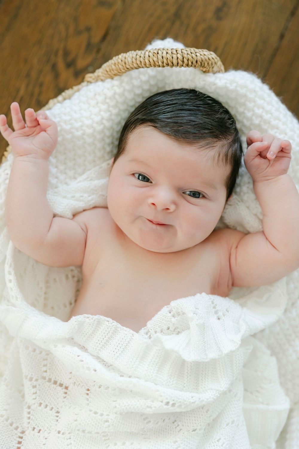 Adorable dark-haired newborn baby lying in wicker basket wrapped in white knit blanket with arms raised.