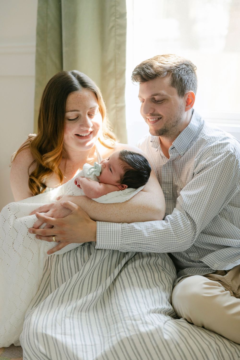 Happy parents smiling at their newborn baby wrapped in a white blanket near a sunlit window.