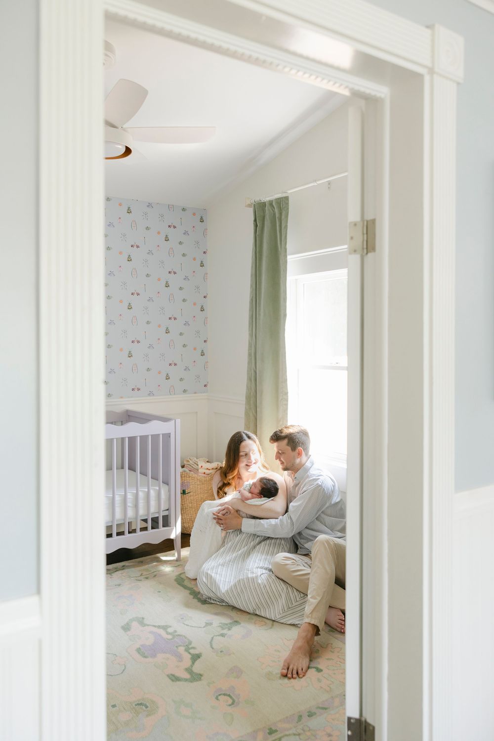 Parents sit on nursery floor with newborn baby, white crib and pastel wallpaper visible through doorway.