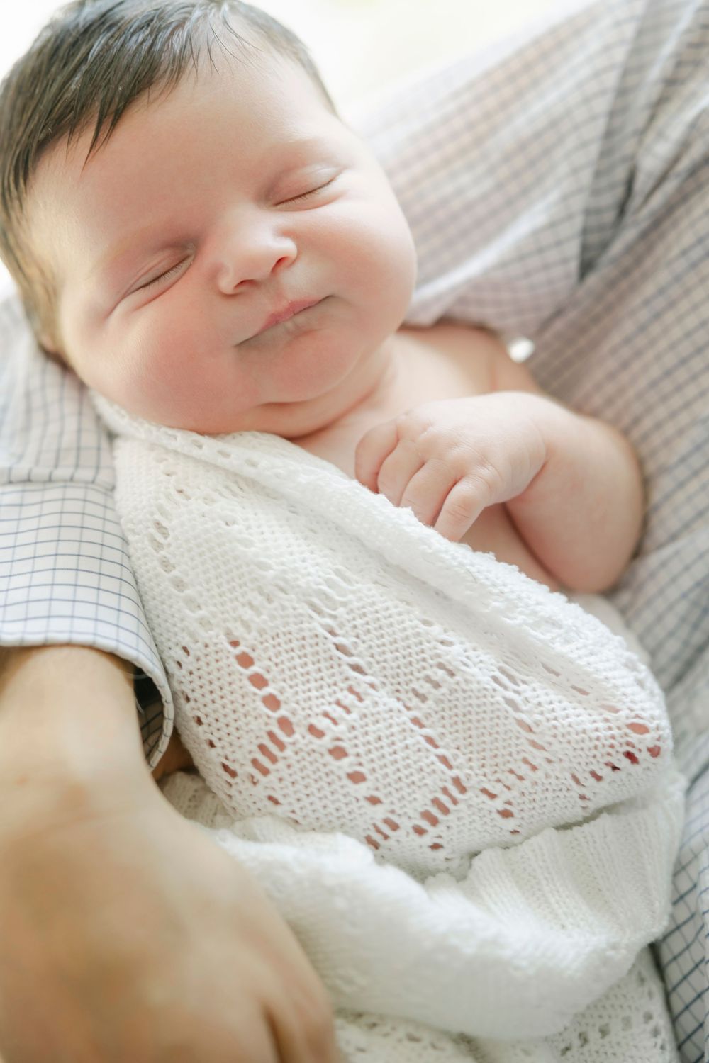 Peaceful newborn baby sleeping wrapped in white knit blanket, cradled in adult arms.