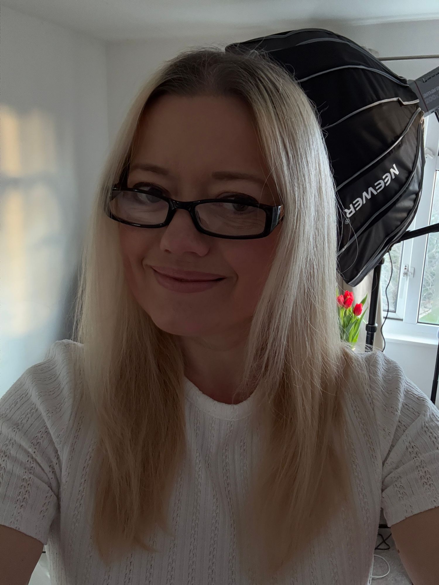 Blonde woman with black glasses smiling, wearing white top, with Neewer softbox photography light in background.
