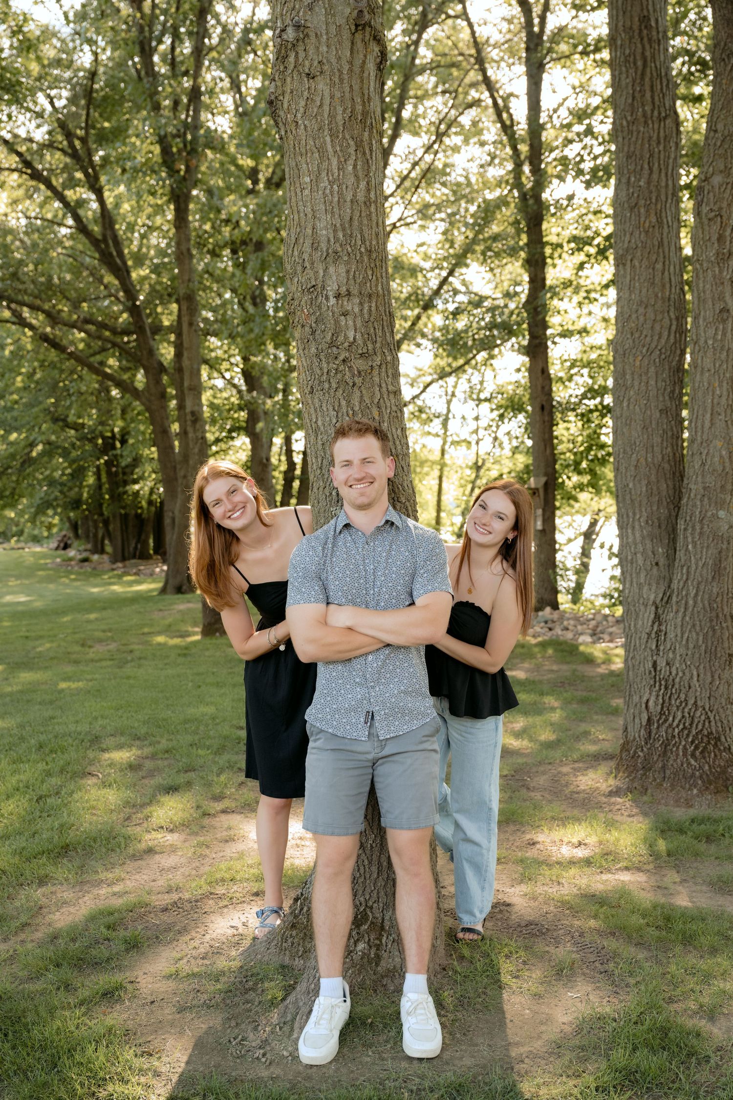 three teenage siblings peek from around a tree for silly family portraits at the cabin on lake wissota in chippewa falls, wi