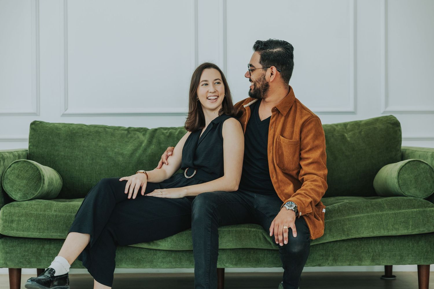 A smiling couple sits together on a green velvet sofa, woman in black dress and man in brown leather jacket.