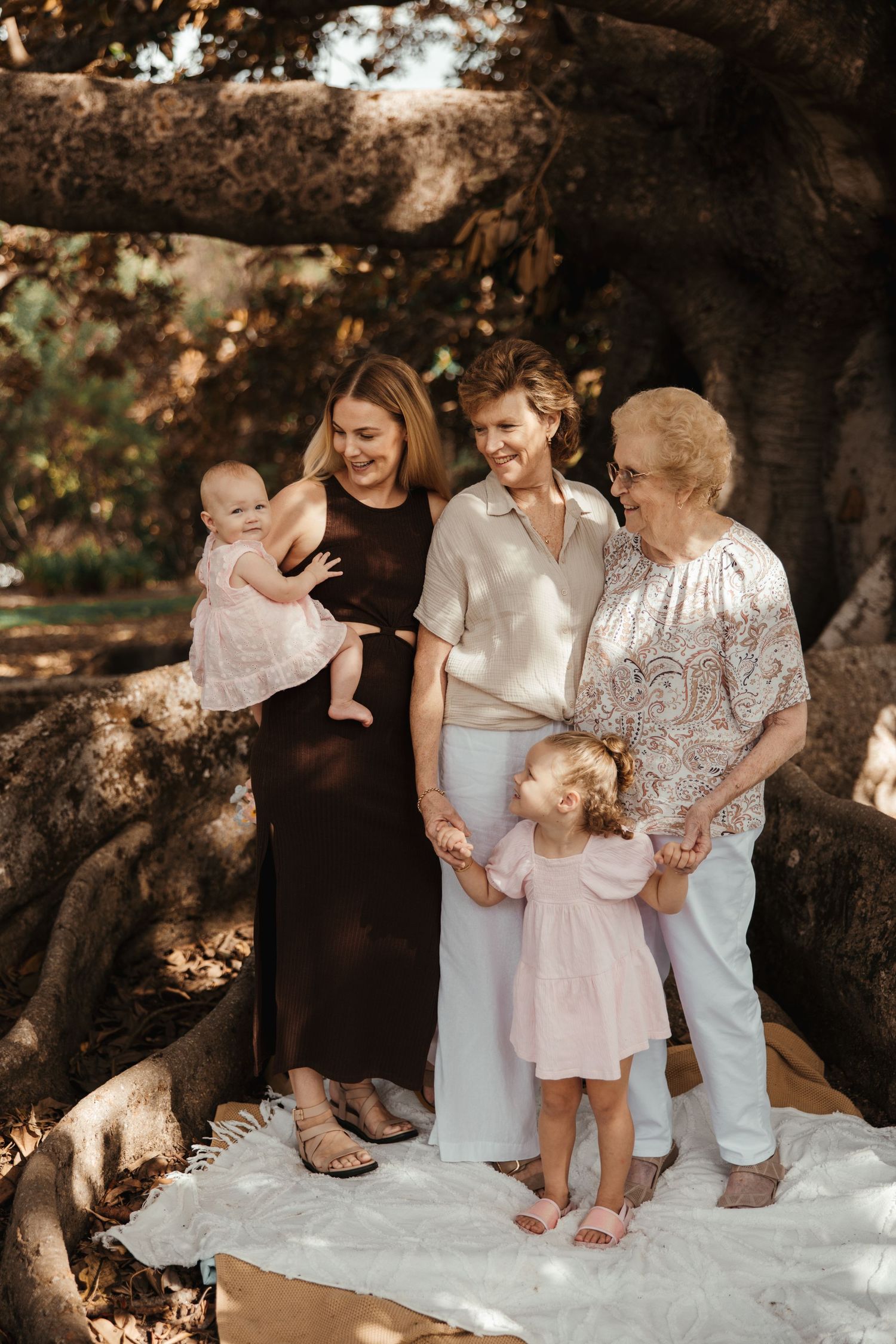 Three generations of women and two young girls pose under a large tree outdoors for a family portrait.