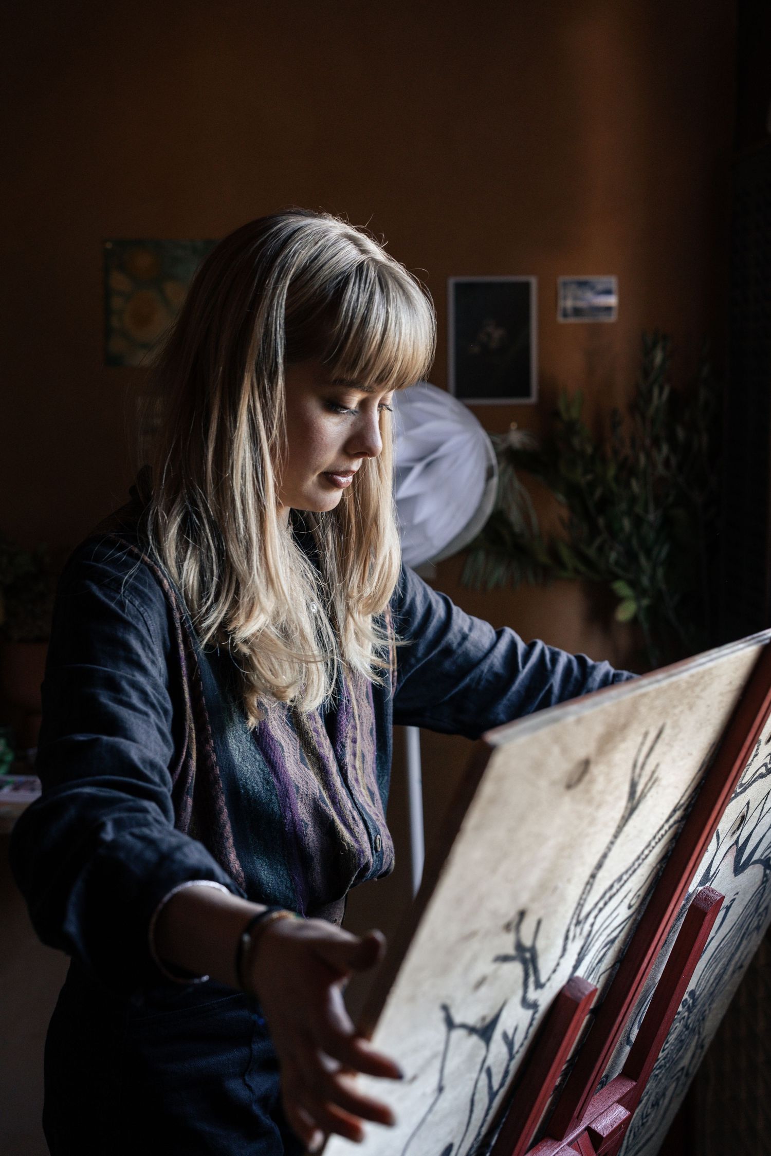 A blonde woman examines a detailed tree artwork on a canvas easel in a dimly lit studio.

Maya Caroll-Tischler