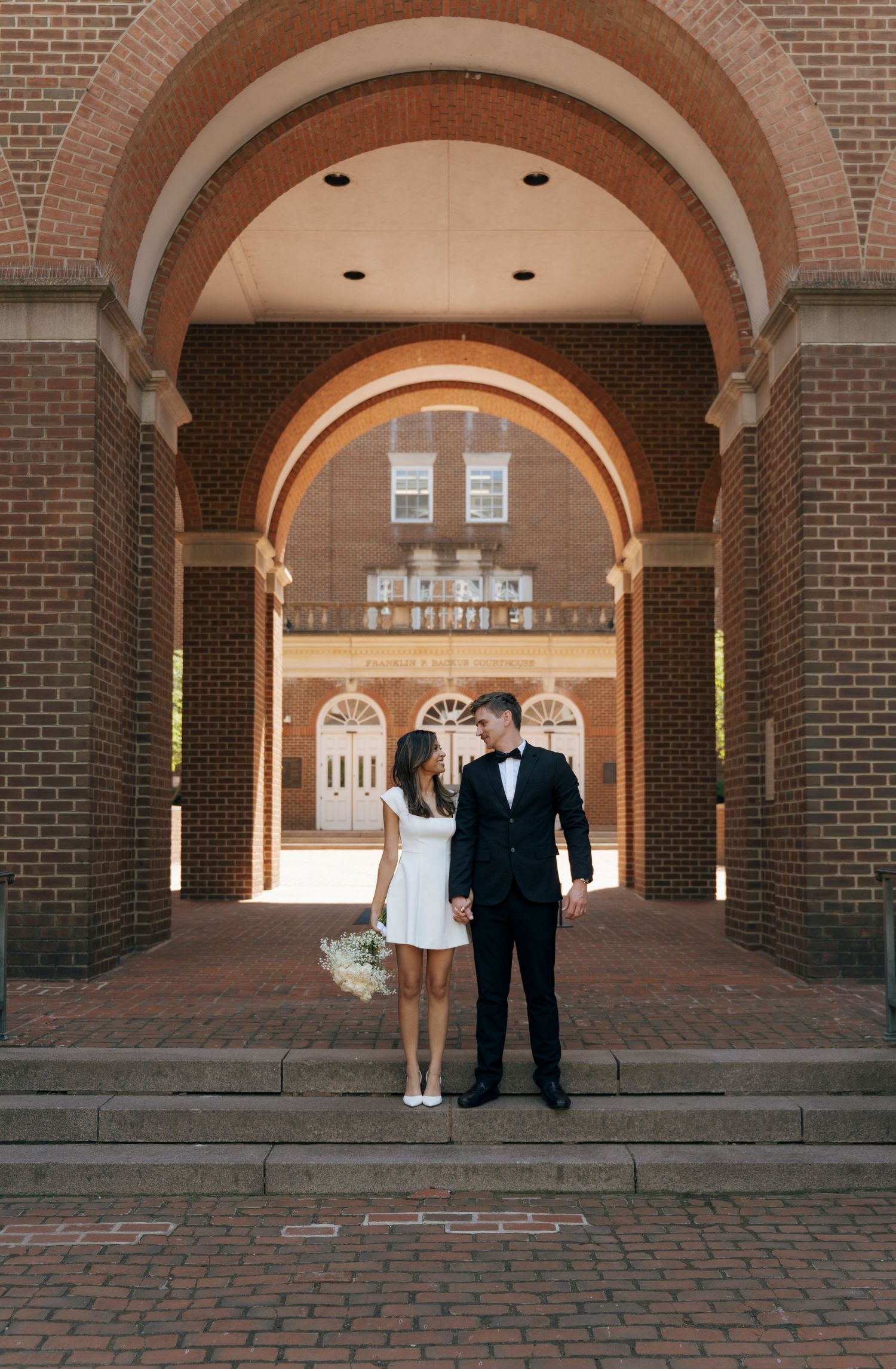 Bride and groom holding hands under a brick archway at an elegant university building on their wedding day.
