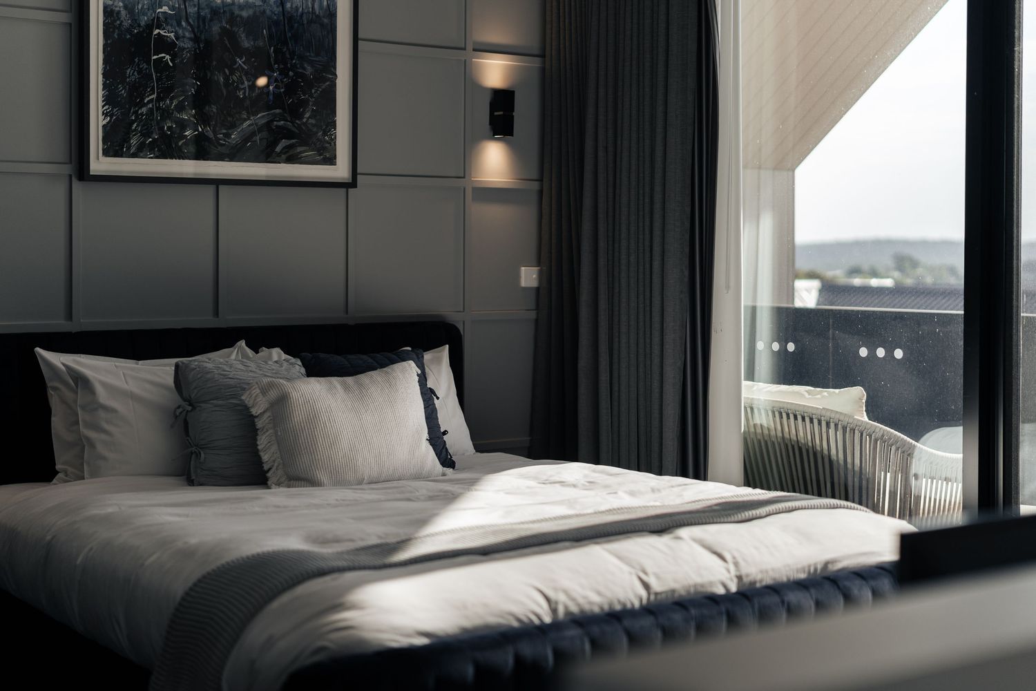 Elegant dark bedroom with upholstered headboard, white bedding, wall sconces, and city view through large floor-to-ceiling window.Lux on Max