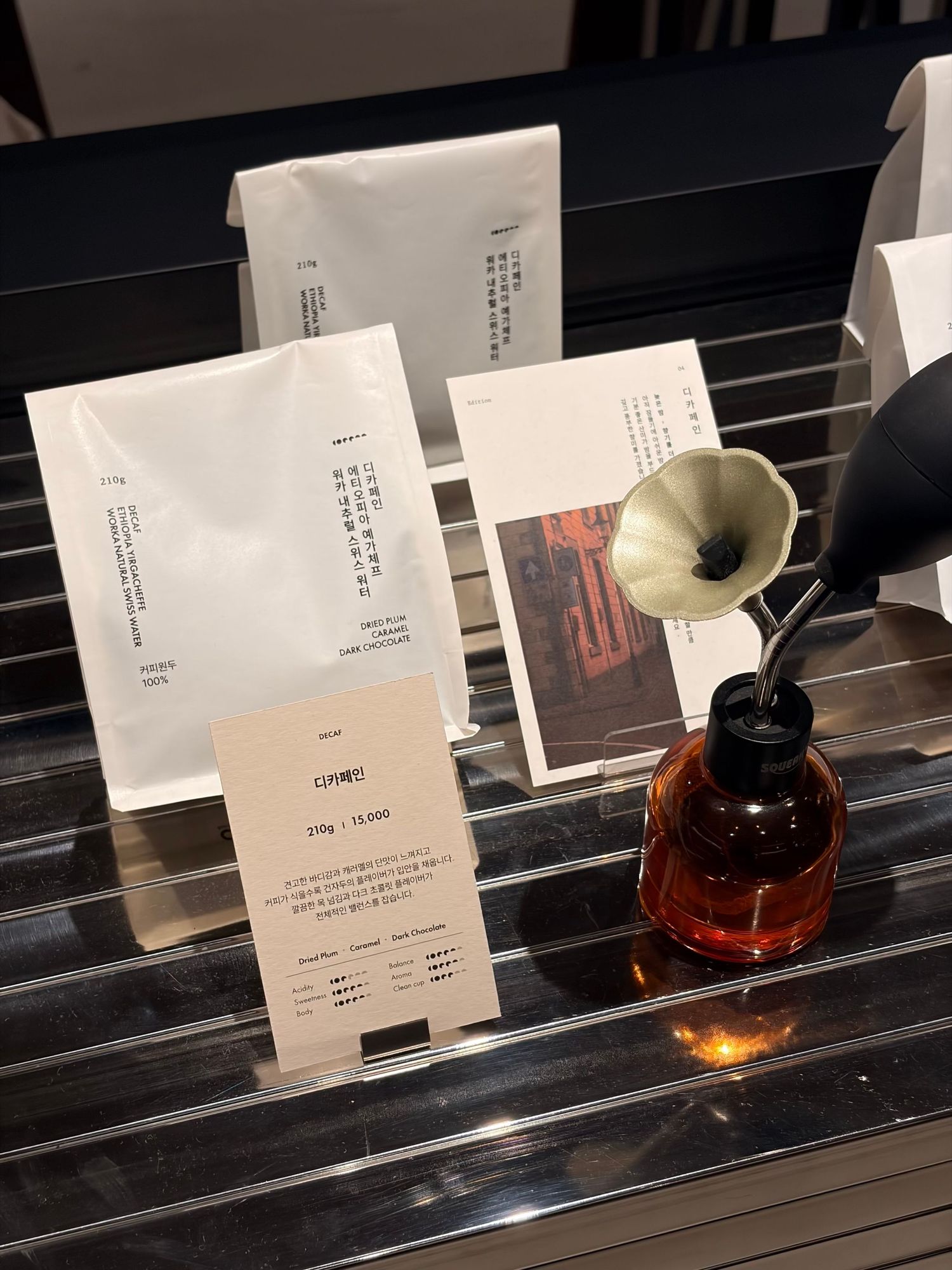 Korean coffee shop display with white packaging bags, price tag, and decorative amber bottle with mini gramophone on black grid surface.