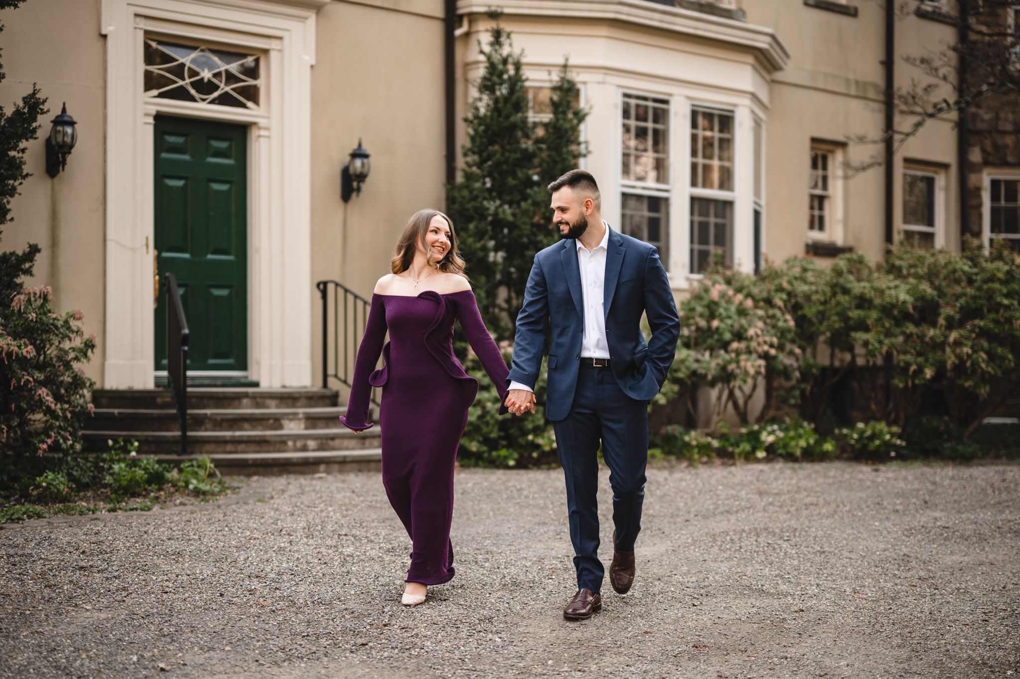 Couple holding hands walking on gravel path outside elegant building, woman in purple gown and man in navy suit.