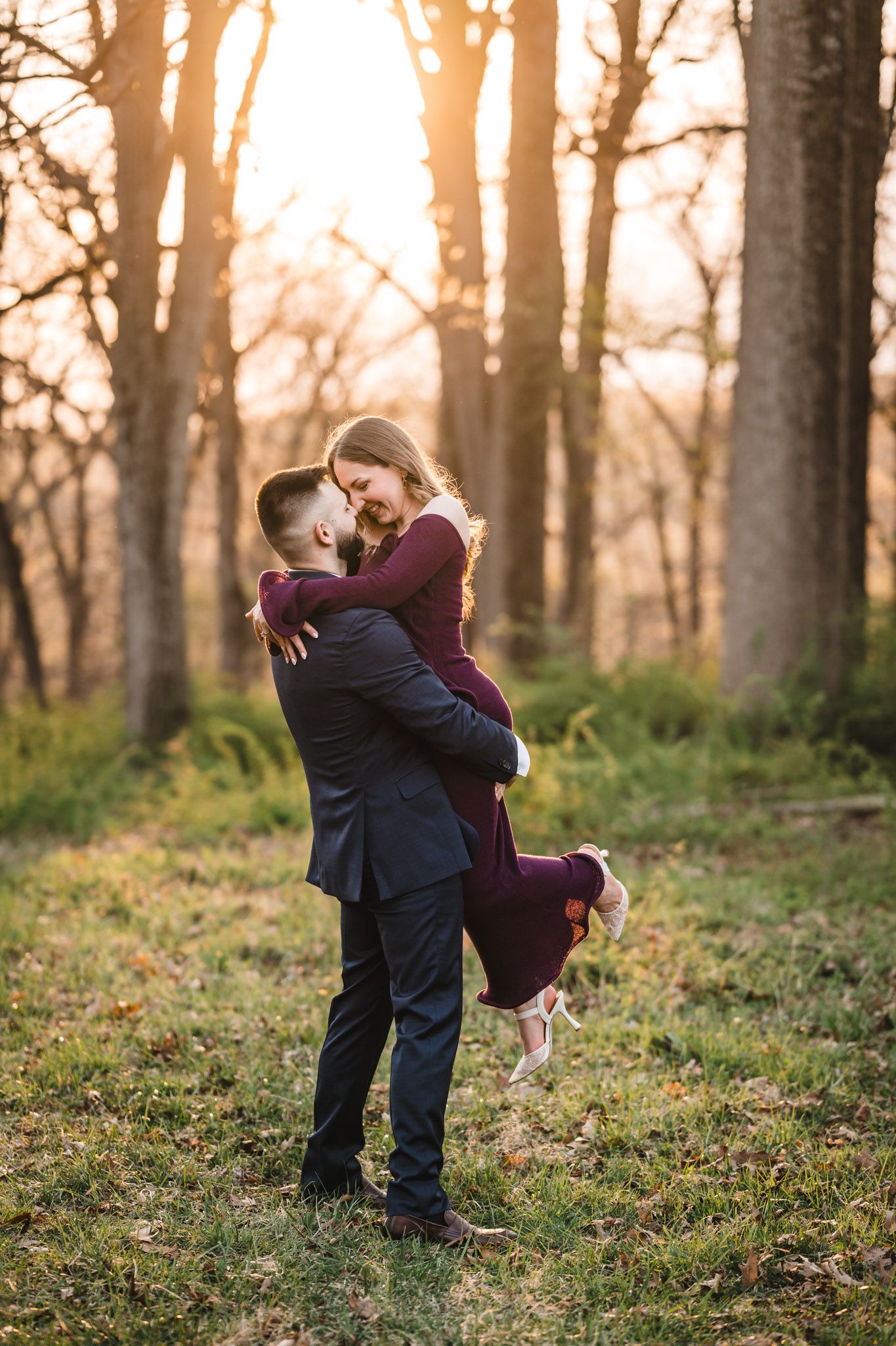A couple shares a romantic embrace at sunset in a forest, man lifting woman in burgundy dress.