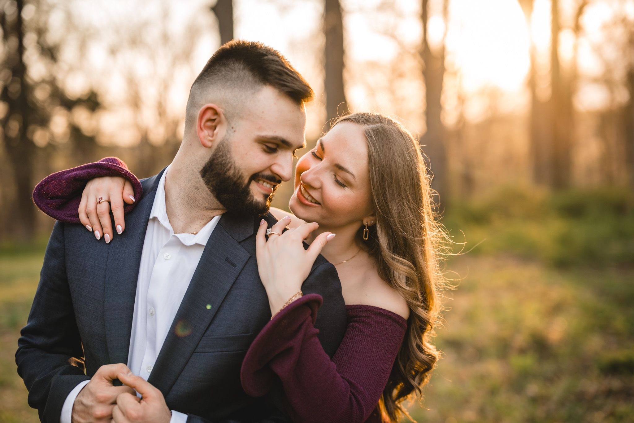 Romantic couple embracing outdoors at golden hour, woman in burgundy off-shoulder top smiling at bearded man in navy blazer.