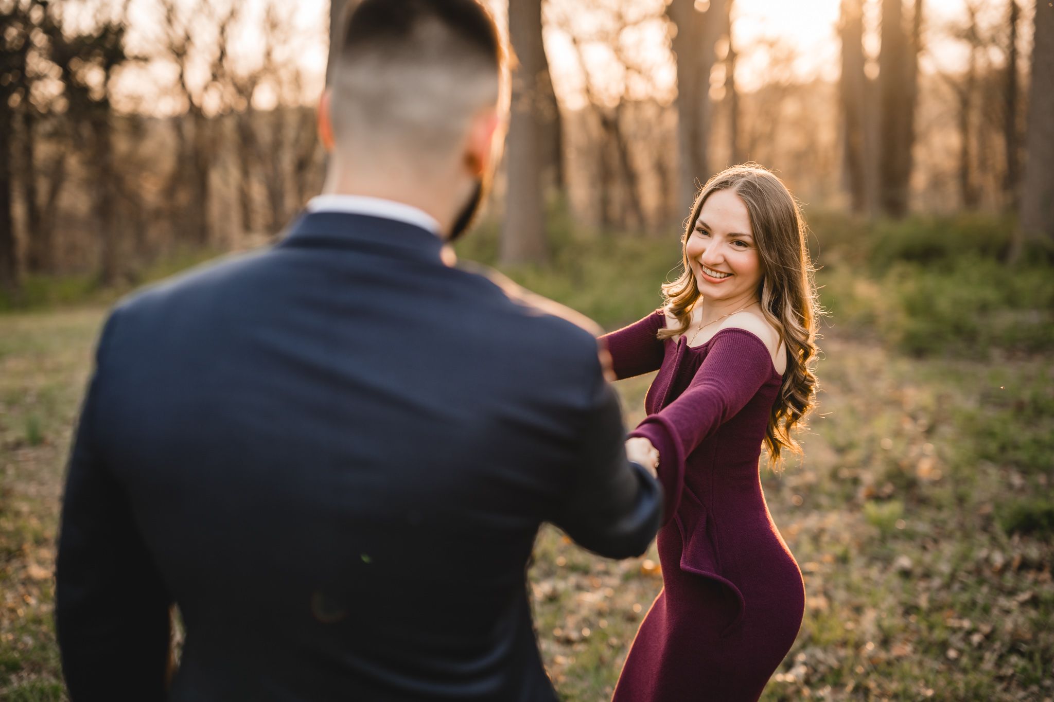 A smiling woman in a burgundy dress holds hands with a man in a navy suit during a golden hour engagement session.