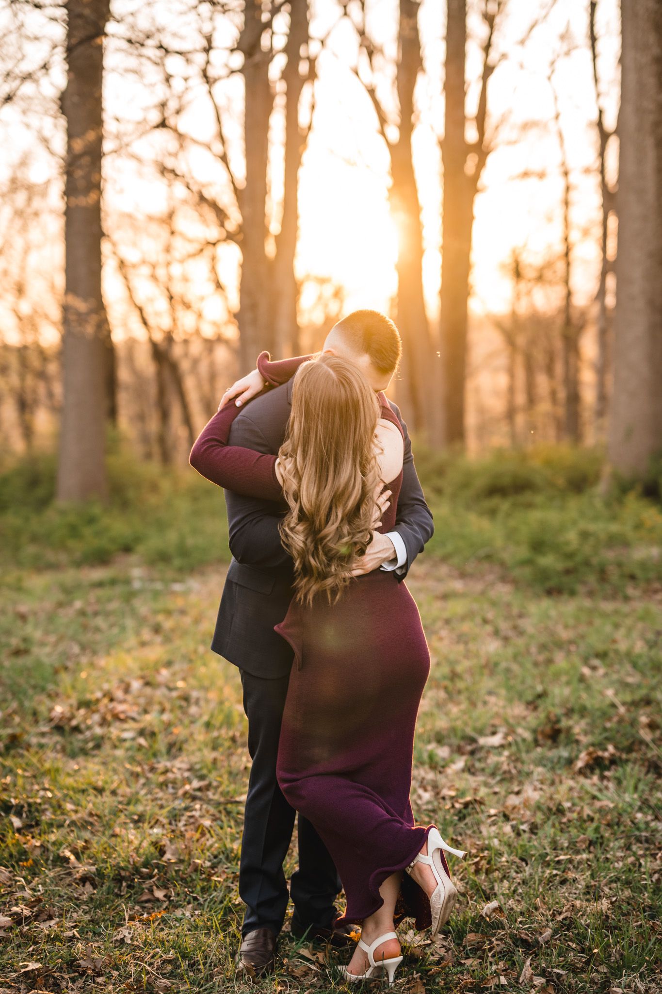 Couple embracing in golden sunset light among bare trees, woman in burgundy dress with flowing blonde hair.