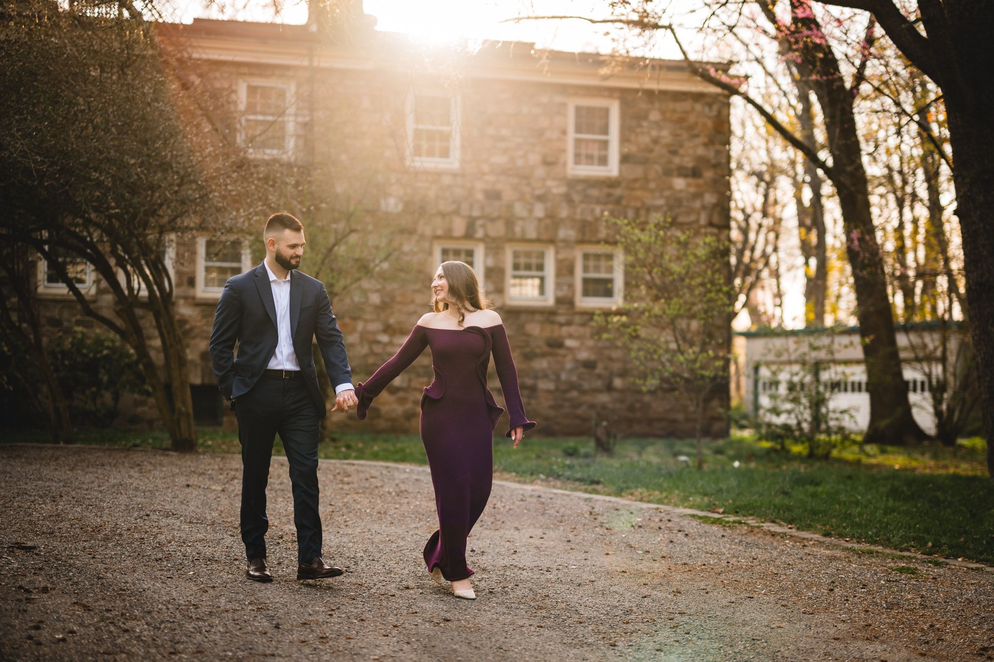 Couple holding hands walking on gravel path at golden hour near historic stone building.