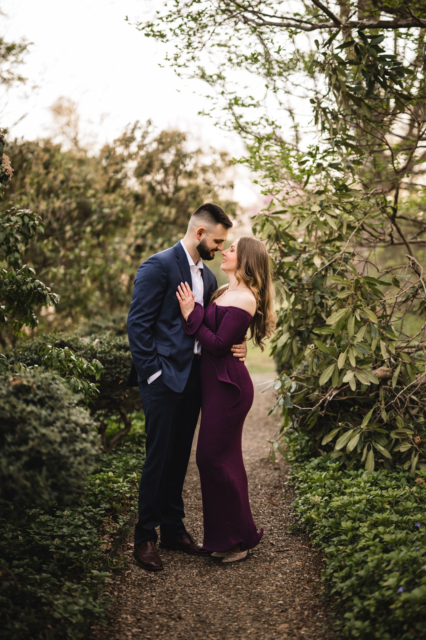 Couple embracing on a garden path, man in navy suit and woman in burgundy off-shoulder gown, surrounded by lush greenery.