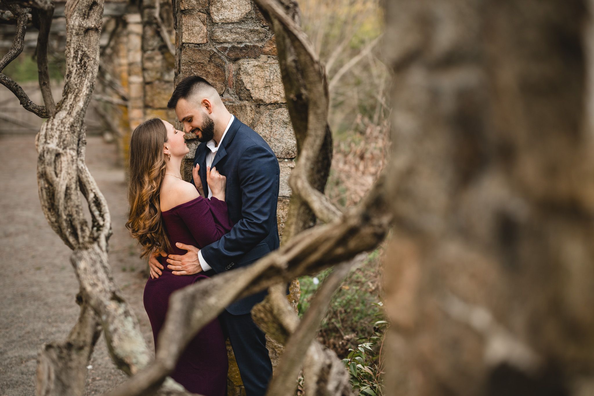 Couple embracing romantically among twisted tree branches with a stone wall backdrop in an outdoor engagement photo.