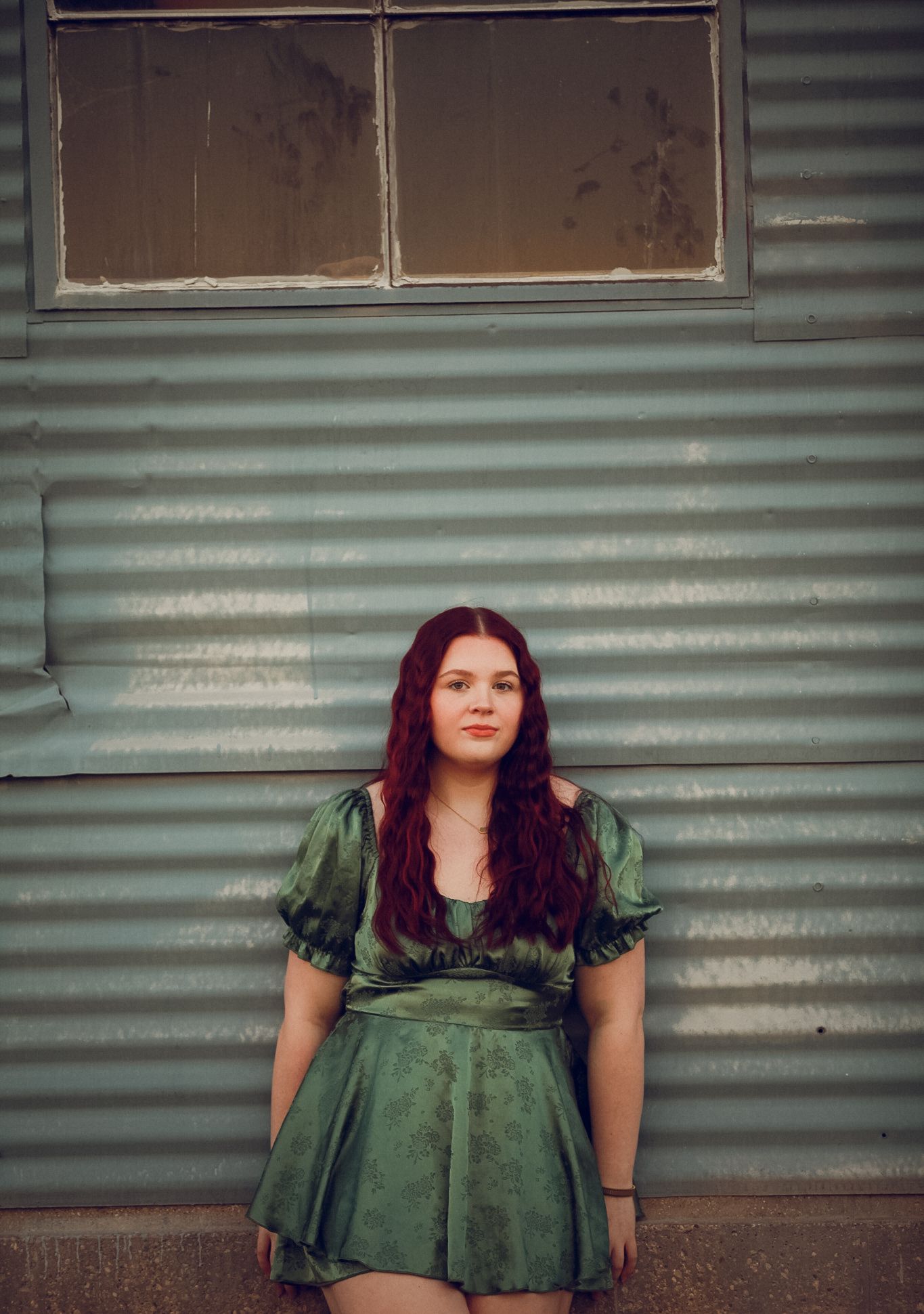 Young woman with auburn hair in a green dress poses against a corrugated metal wall outdoors.