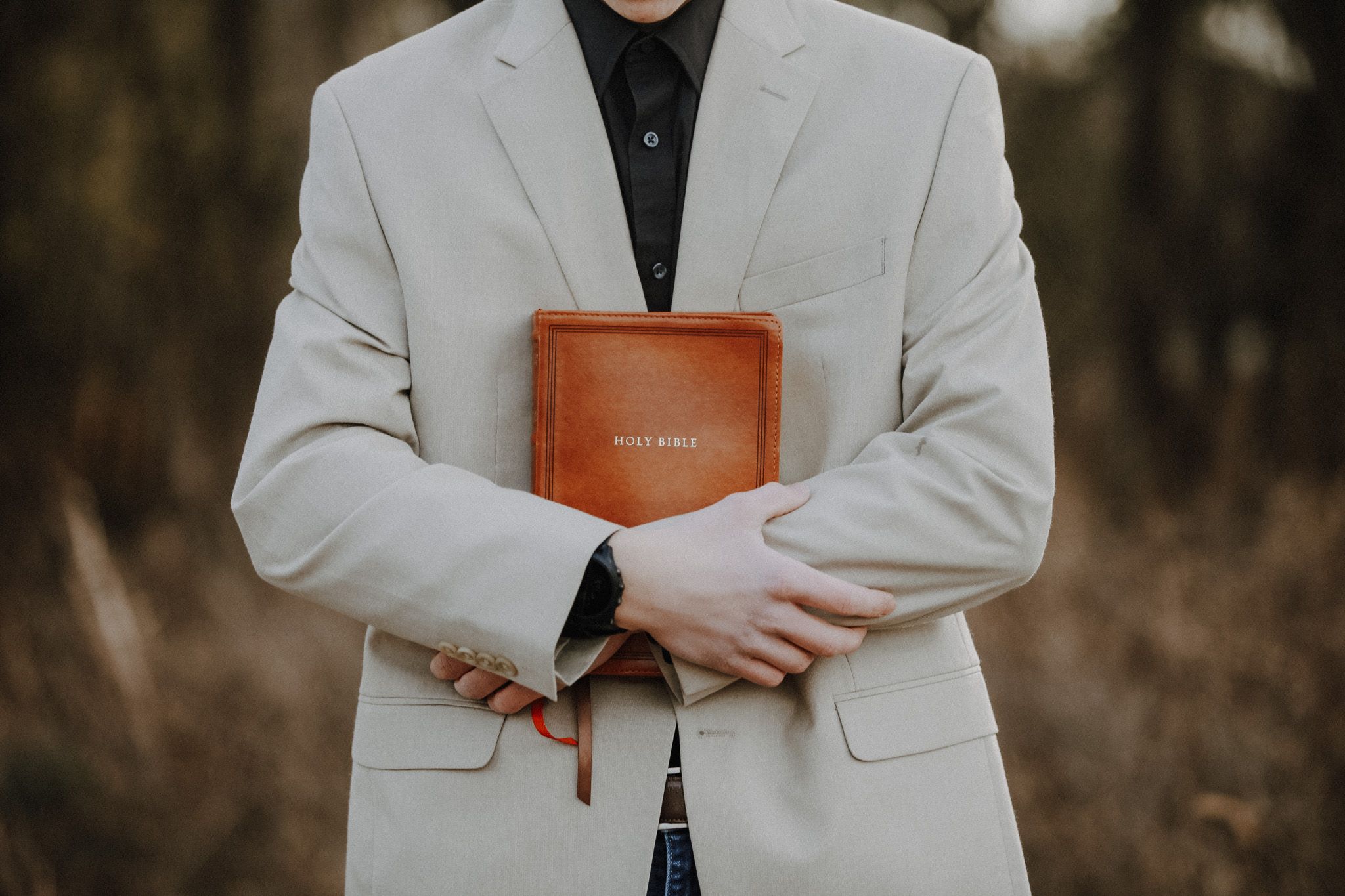 Person in beige blazer holding a brown Holy Bible outdoors in a natural setting.